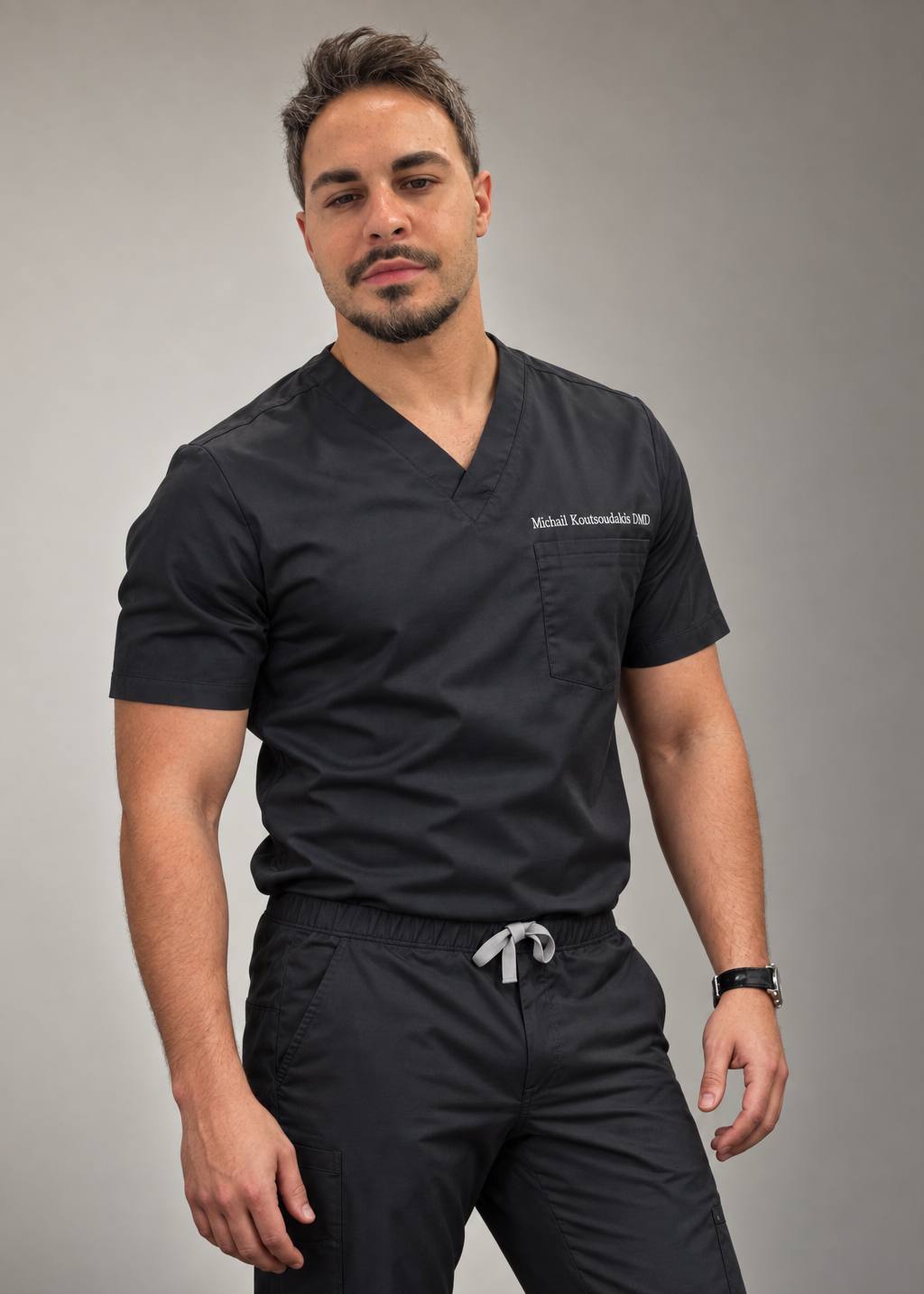 Michail Koutsoudakis standing, in black scrubs embroidered with his name, against a neutral grey backdrop